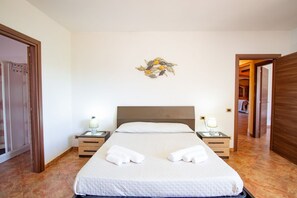 Een netjes opgemaakt bed met witte lakens, twee nachtkastjes met lampen en een aan de muur hangend kunstwerk.