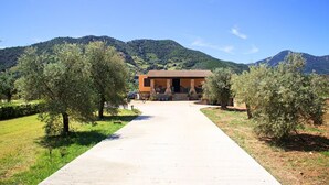 Uma casa com telhado de cerâmica, rodeada por oliveiras e uma montanha ao fundo.