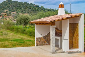 Uma pequena cozinha ao ar livre com parede de pedra e telhado ladrilhado, situada em uma encosta com casas ao fundo.