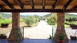 Área externa coberta com pilares de pedra e piso de azulejos, que dá acesso a um caminho por um jardim paisagístico.