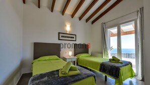 Une chambre d’hôtel avec deux lits, une table de chevet, une lampe fixée au mur et une vue sur la mer grâce à de grandes fenêtres.