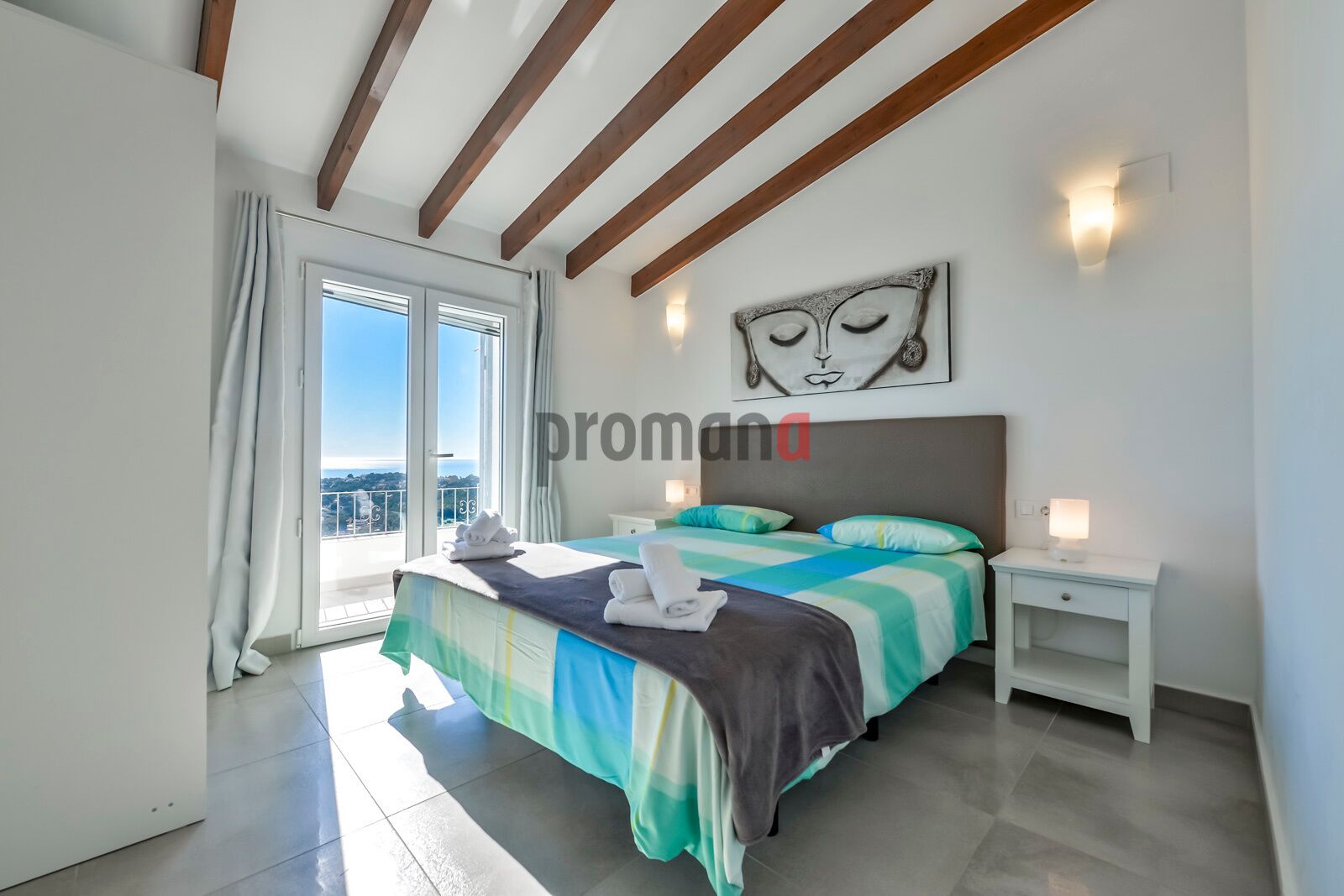 Een slaapkamer met een bed, nachtkastjes, een schilderij en uitzicht door een groot raam.