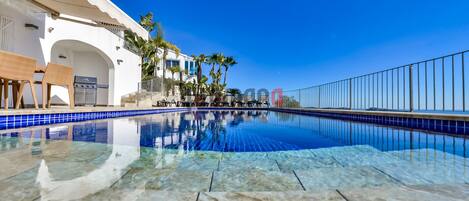 A clear blue pool with tiled edges, surrounded by lounge chairs and a white building with balconies.