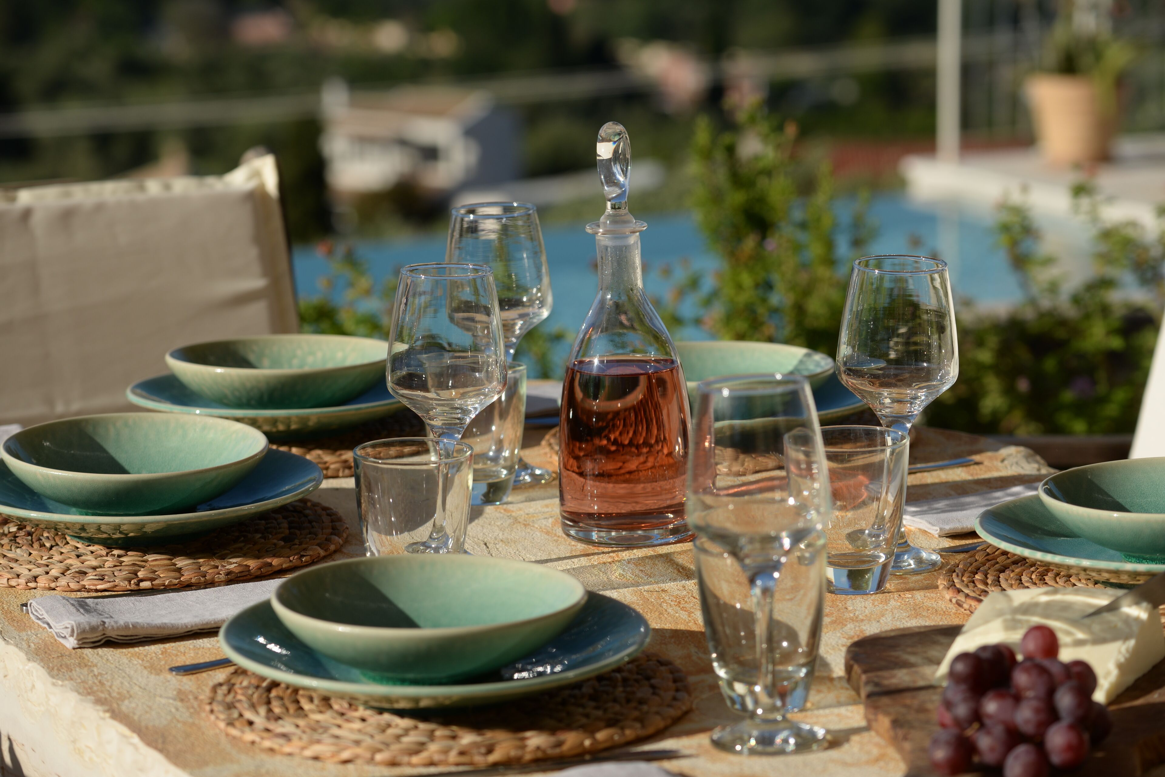 A table set with green plates, clear wine glasses, a decanter, and grapes, overlooking a body of water.