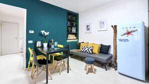 A modern living space with a dining area, a sofa with cushions, a small stool, a refrigerator, and a bookshelf against a teal wall. - Design seashell design (San Pellegrino terme bergamo)
