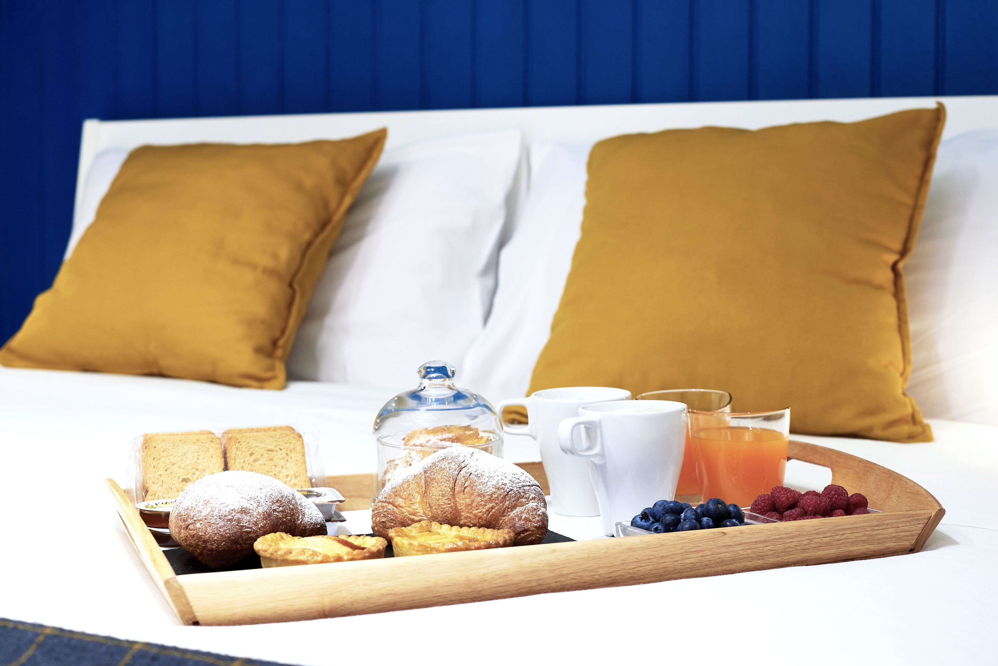 Un vassoio colazione con pasticcini, frutta e bevande su un letto con biancheria bianca e cuscini color senape.
