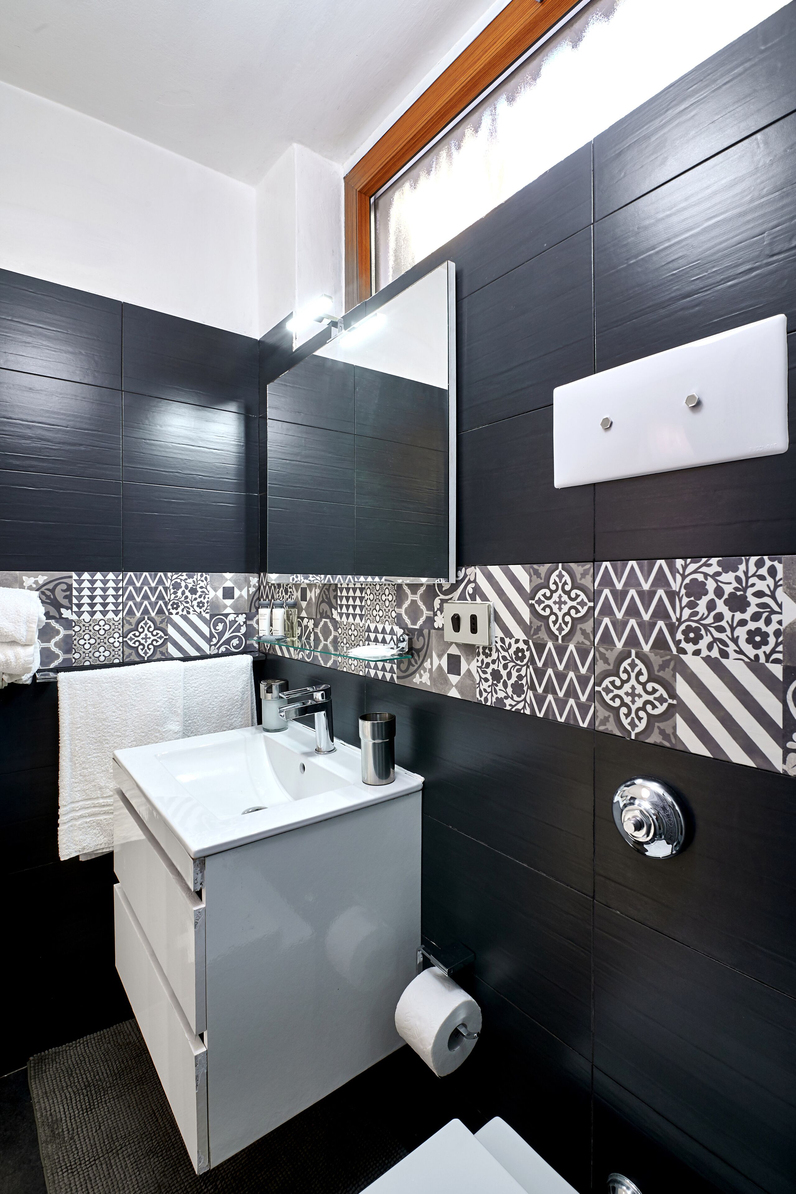 Une salle de bain moderne avec un lavabo blanc, des murs recouverts de carreaux foncés, un miroir et un porte-serviettes.