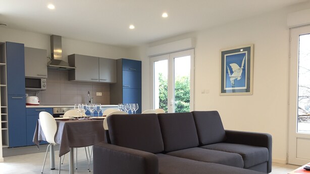 A modern living room with a dark sofa, a dining area with a table and chairs, and a kitchen with blue cabinets and a microwave. - 3-bedroom house near Colmar (Bennwihr)