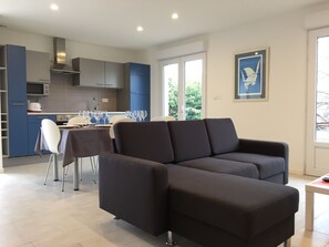 Ein modernes Wohnzimmer mit einem dunkelgrauen Sofa, einem Essbereich mit Tisch und Stühlen sowie einer Küche mit blauen Schränken und einer Mikrowelle.