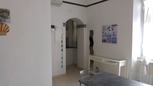 Una habitación con pared blanca, una puerta que da a otra estancia, una pequeña mesa con una silla y un estante fijado a la pared.