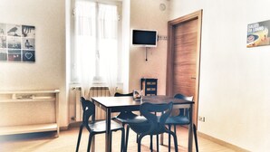 Un comedor con una mesa y sillas de madera, un televisor montado en la pared y una ventana con cortinas ligeras.