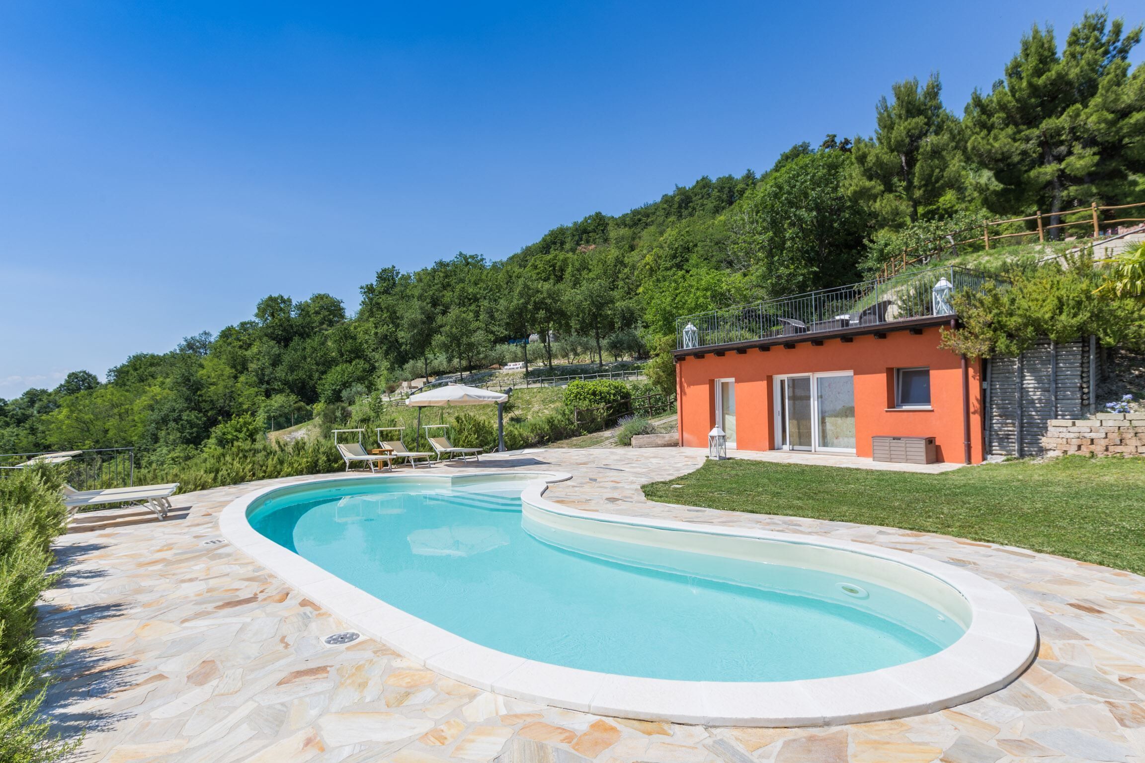 A swimming pool with a stone deck, a small building with a balcony, and a hillside with trees.