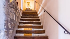 Un escalier en bois, avec un mur rustique d’un côté et un mur blanc de l’autre, mène à une pièce où se trouvent une lampe et un tableau encadré.
