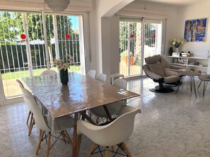 A dining area with a table, chairs, a vase with flowers, a sofa, and a bookshelf.