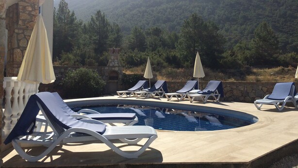 A pool area with lounge chairs and umbrellas, surrounded by stone walls and a view of a forested mountain.
