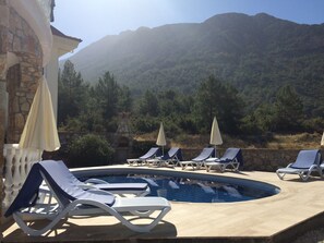 A pool area with lounge chairs and umbrellas, surrounded by stone walls and a view of a forested mountain. - Detached Villa - Private Pool - Stunning Mountain Views (12-2 Bn. B Dr. Fethiye)
