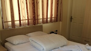 A neatly made bed with white linens and towels, a bedside table with a lamp, and a window with patterned curtains.