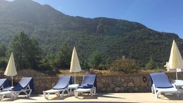 A poolside area with lounge chairs and umbrellas, a stone wall, and a mountainous backdrop.