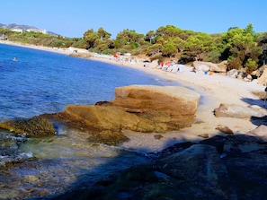 Una playa con agua cristalina, un promontorio rocoso y una hilera de árboles al fondo.