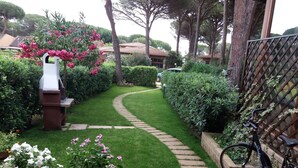 Un sentier de jardin mène à une maison dotée d’un espace barbecue, le tout entouré d’une végétation luxuriante, avec un vélo garé près de la clôture.