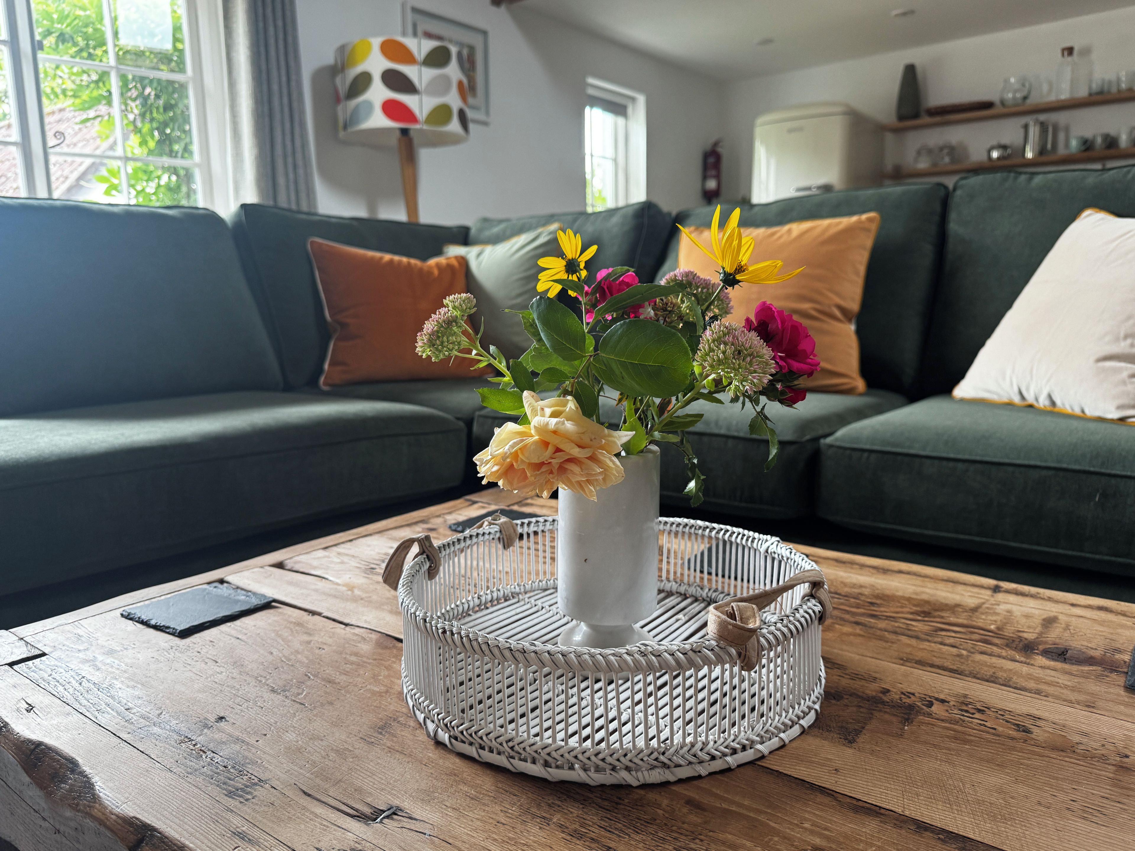Een woonkamer met een bank, een bijzettafeltje met een geweven dienblad, en een vaas met bloemen.