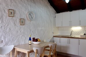 Een keuken met witte kastjes, een houten tafel en stoelen. Er hangt een klok aan de muur en op de tafel staat een schaal fruit.