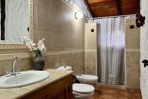 Un baño con lavamanos, dos inodoros, un bidé, ducha con azulejos estampados y un espejo con un florero.