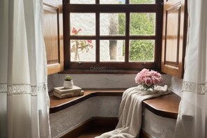 Una ventana de esquina con vistas a follaje, una pequeña planta en maceta y un florero sobre un repisa de madera.