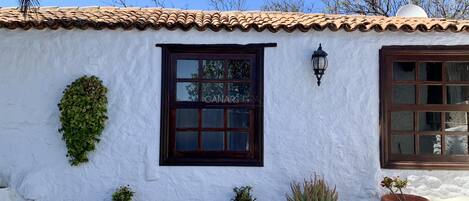A building with a white stucco exterior, terracotta roof tiles, and a window with a dark frame. There are potted plants on the ground.