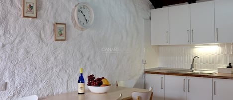 Una cocina con armarios blancos, una mesa de madera y sillas. Hay un reloj en la pared y un cuenco con fruta sobre la mesa.