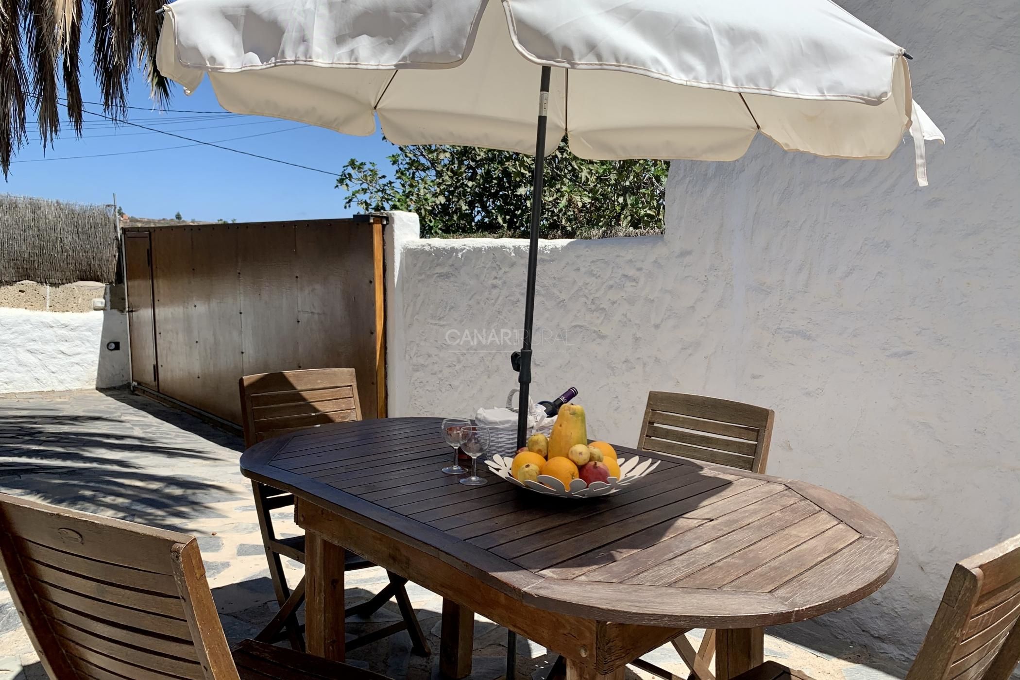 Uma mesa de madeira com cadeiras, um guarda-sol branco e uma cesta de frutas.