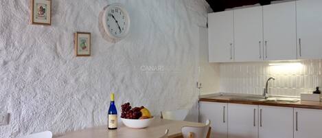 A kitchen with white cabinets, a wooden table, and chairs. There is a clock on the wall and a bowl of fruit on the table.