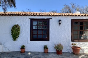 Een gebouw met een witte stucgevel, terracotta dakpannen en een raam met donkere omlijsting. Er staan bloempotten op de grond.