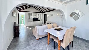 A dining area with a table and chairs, a kitchen in the background, and a hallway leading to an outdoor area.