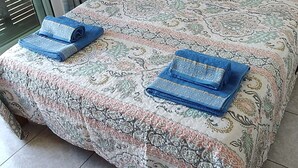 Una cama bien hecha con una colcha estampada, dos toallas enrolladas, un cabecero de madera, una mesita de noche con una lámpara y una ventana con cortinas.