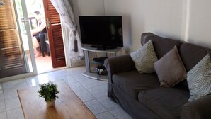 Un salón con una mesa de centro de madera, un sofá con cojines estampados, un televisor de pantalla plana sobre un mueble, y un ventanal corredero que da a un espacio exterior.