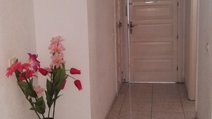 Un pasillo con suelo de baldosas y una puerta al final. Se ve un jarrón con flores en la pared izquierda.