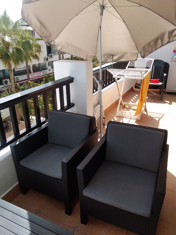 Un balcon avec deux fauteuils en osier de couleur sombre, un grand parasol beige et une table blanche.