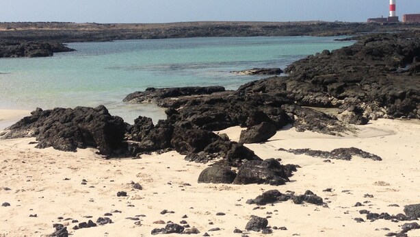 Ein Strand mit felsigem Gelände und einem Leuchtturm in der Ferne.