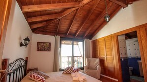 Ein Schlafzimmer mit Holzboden, einem Bett mit gemusterten Kissen, einem Stuhl und Blick ins Badezimmer.
