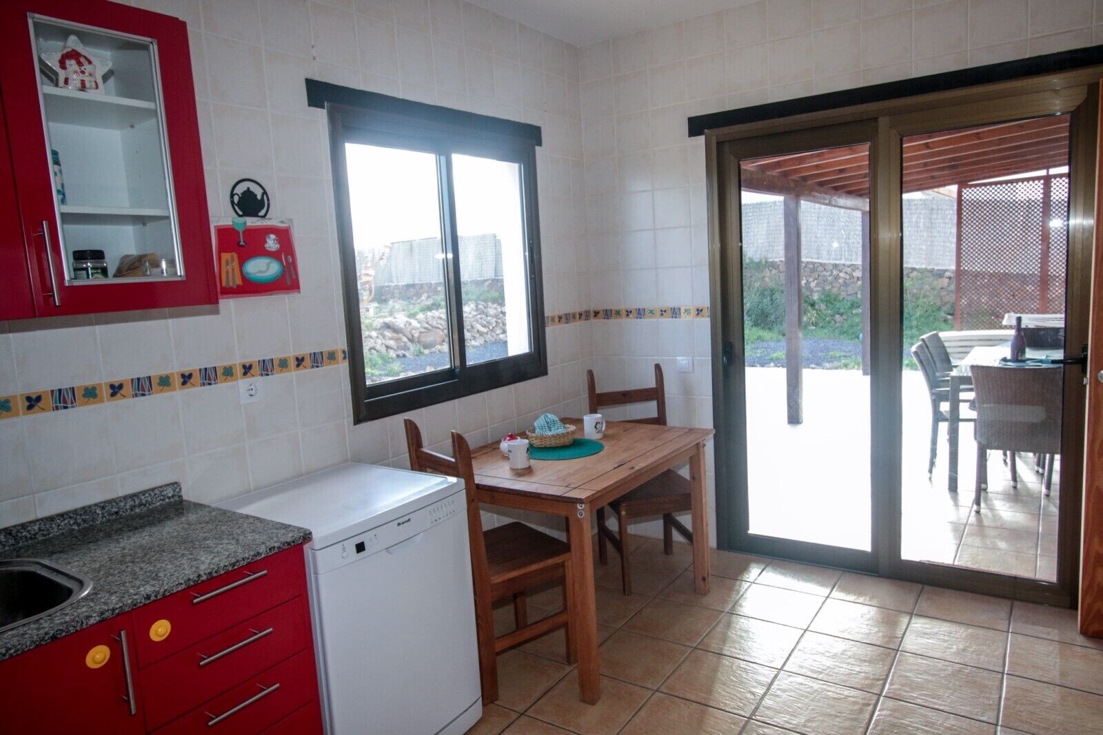 Una cocina con un armario rojo, un lavavajillas blanco y una mesa de comedor con sillas. Hay una ventana que ofrece vistas al exterior.