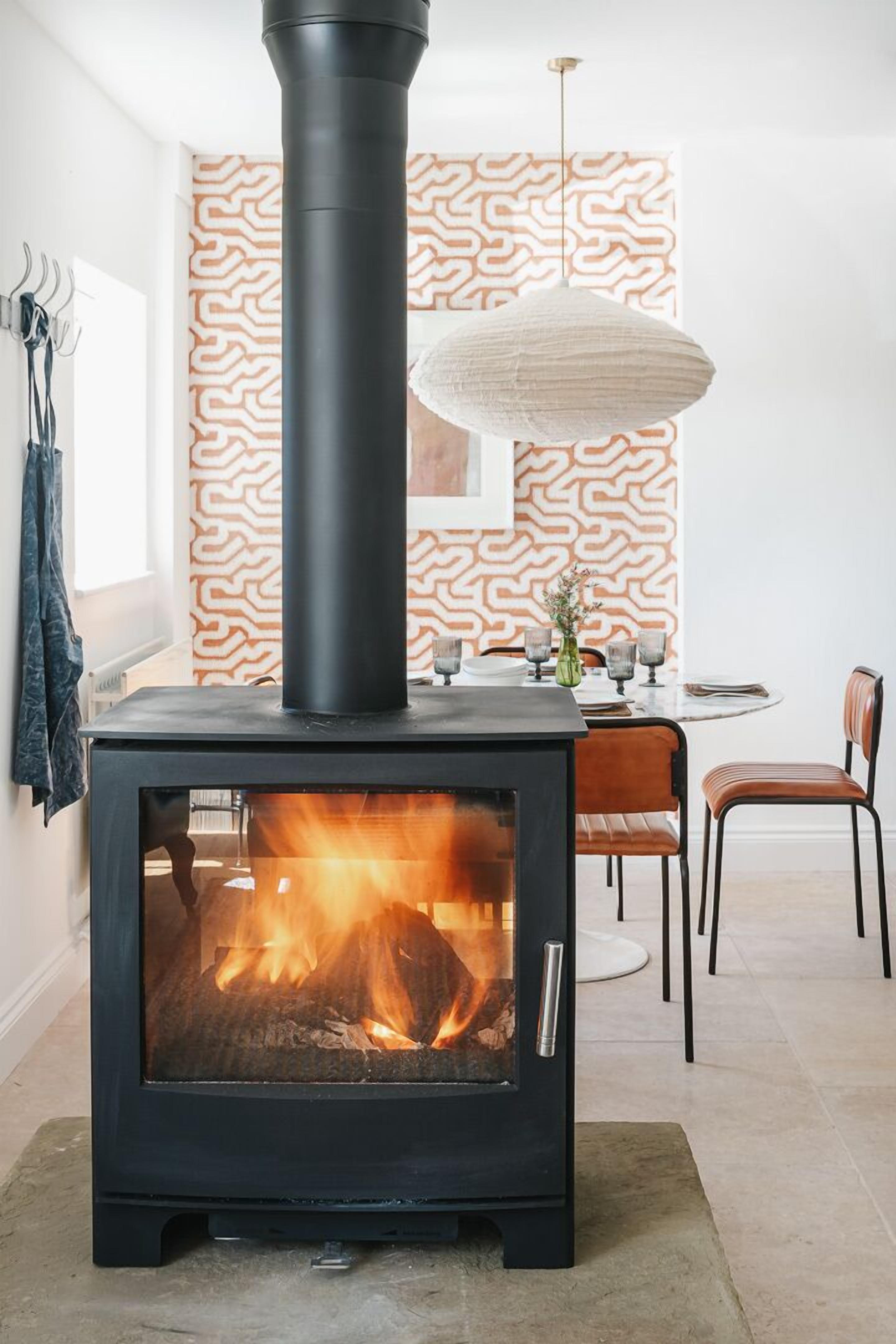 Ein modernes Wohnzimmer mit einem schwarzen Kaminofen, einer Wand mit Muster, einem Esstisch mit Stühlen und einer hängenden Leuchte.