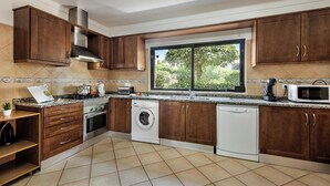 A modern kitchen with wooden cabinets, a built-in stove, a washing machine, and a large window offering a view of greenery. - Exclusive Retreat in Vilamoura, private pool, off-street parking, Free AC & wifi (Vilamoura)