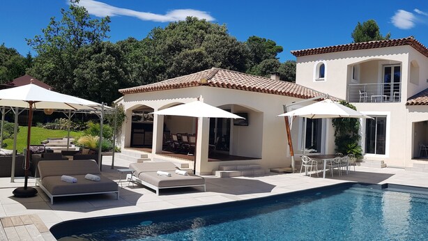 Zona junto a la piscina con sillones, sombrillas y un edificio de dos plantas con techo de tejas.
