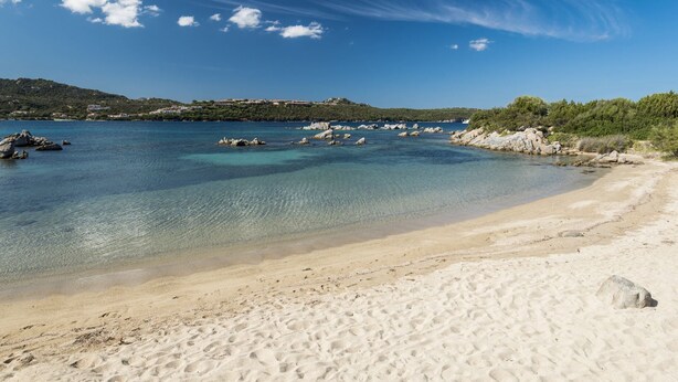 A sandy beach with clear water, rocky outcrops, and a distant hill with buildings. - Villaggio S'abba e Sa Pedra - S'ABBA DI SU (Golfo Aranci)