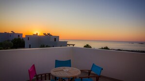 Un balcone con un tavolo rotondo in legno e sedie blu, con vista su un tramonto sul mare e edifici sullo sfondo.
