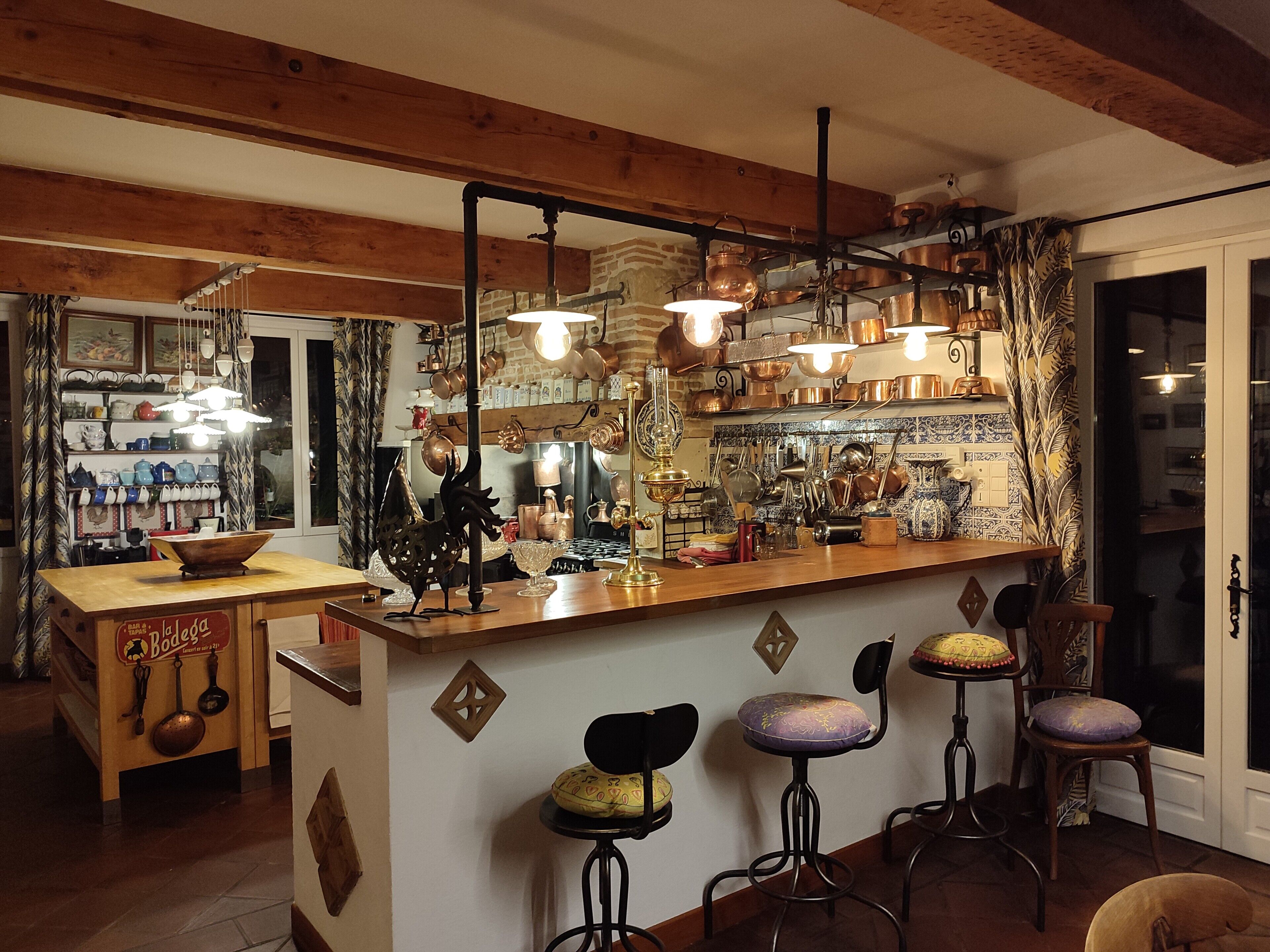 Un coin bar chaleureux avec des poutres en bois, des luminaires suspendus et une variété d’ustensiles de cuisine exposés sur des étagères.