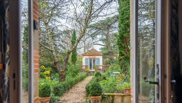 A view through a glass door of a garden leading to a small building with a tiled roof and shutters. - Beautiful house with swimming pool, in Little Tuscany between Albi and Toulouse (Ambres)