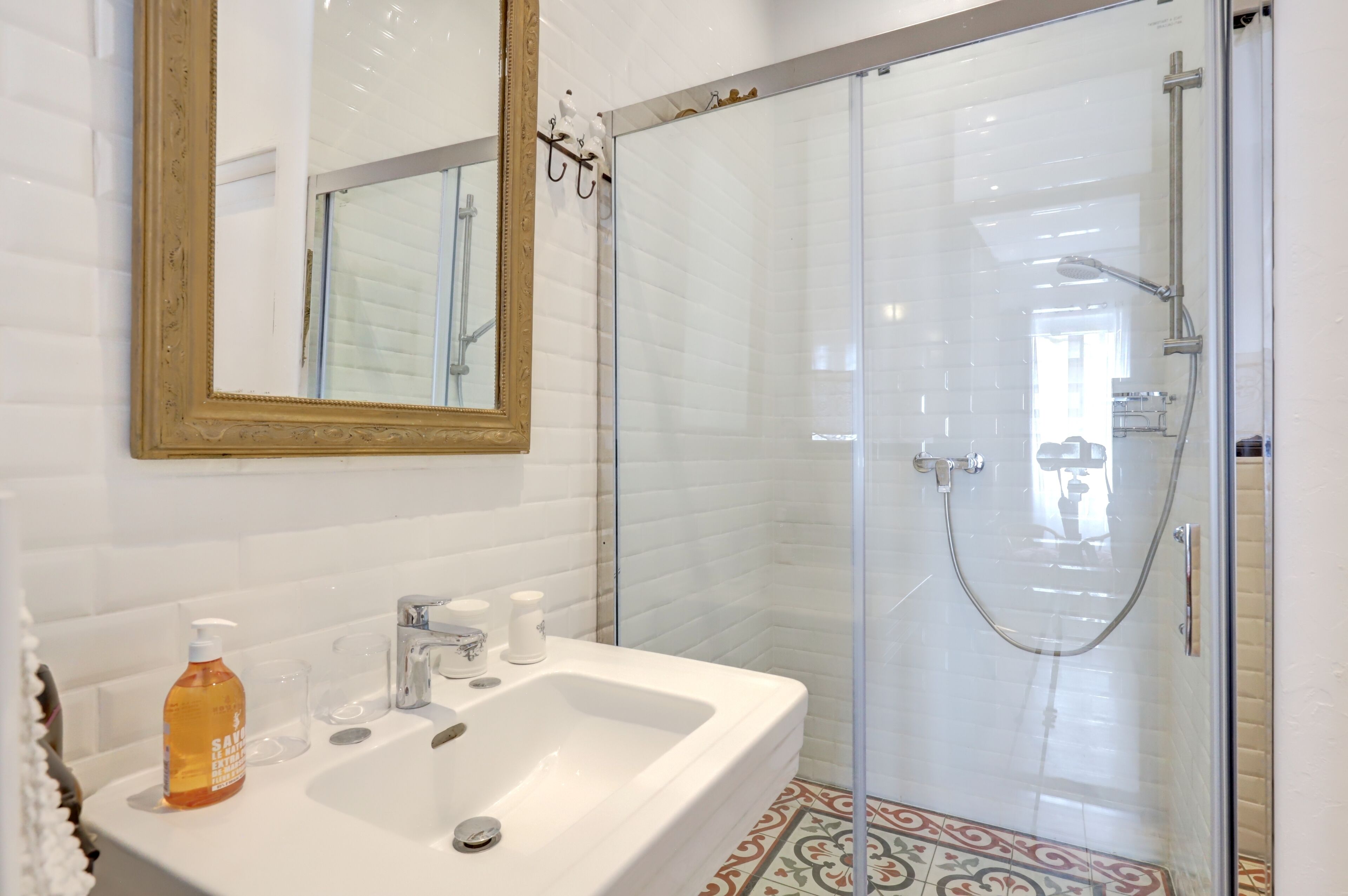Une salle de bain avec un lavabo blanc, un miroir, une douche avec une porte en verre et des carreaux de sol à motifs.