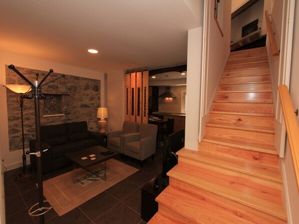 A modern living room with a wooden staircase, a stone accent wall, and a dining area.